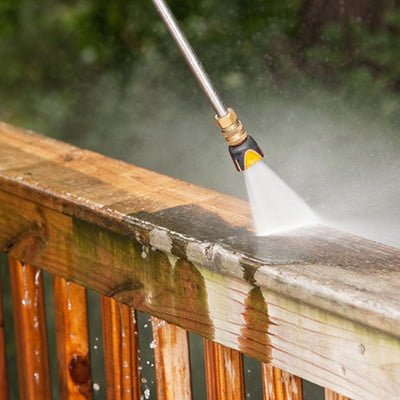 A pressure washer is being used for home improvement, with a focused spray of water cleaning a wooden deck railing and exposing the clean wood beneath.