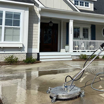 A driveway cleaning surface cleaner is used on a wet concrete driveway in front of a house undergoing a cleaning service