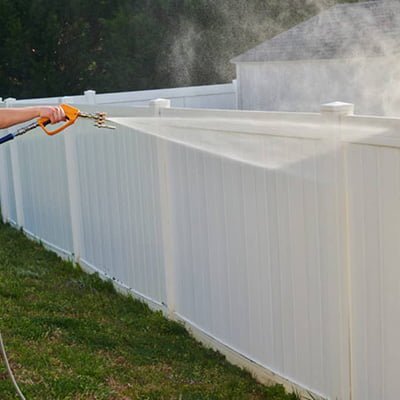 A person pressure washing a white vinyl fence, effectively removing dirt and grime as part of a fence restoration project, with visible spray and clean section on the fence.