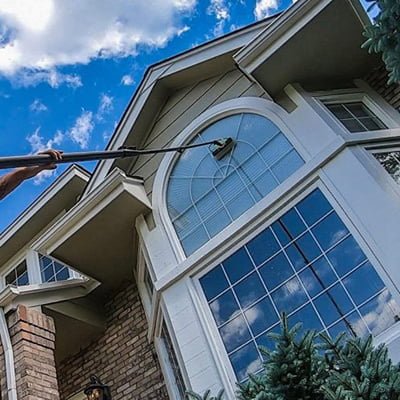 A person using a long-handled squeegee to clean a large arched window on the second story of a residential home, focusing on window washing, with a clear blue sky and fluffy clouds visible